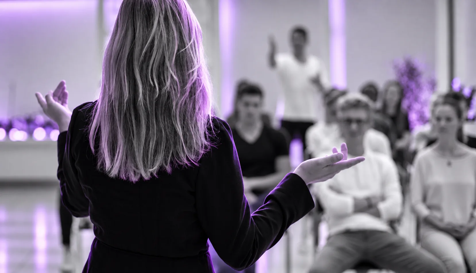 Referentin steht mit ausgebreiteten Armen vor einer Gruppe von Teilnehmenden in einem violett beleuchteten Raum