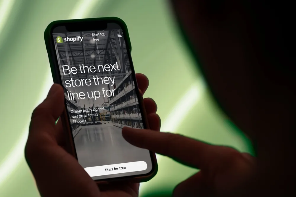 Hand hält ein Smartphone mit der Shopify-Startseite vor grünem Hintergrund
